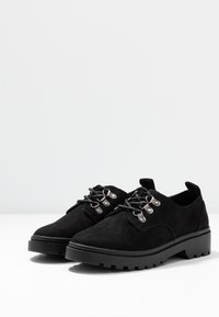 Chaussures en suède noir à lacets avec une semelle chunky, dotées d'œillets en argent et de lacets noirs contrastants. Texture lisse et design à bout rond.