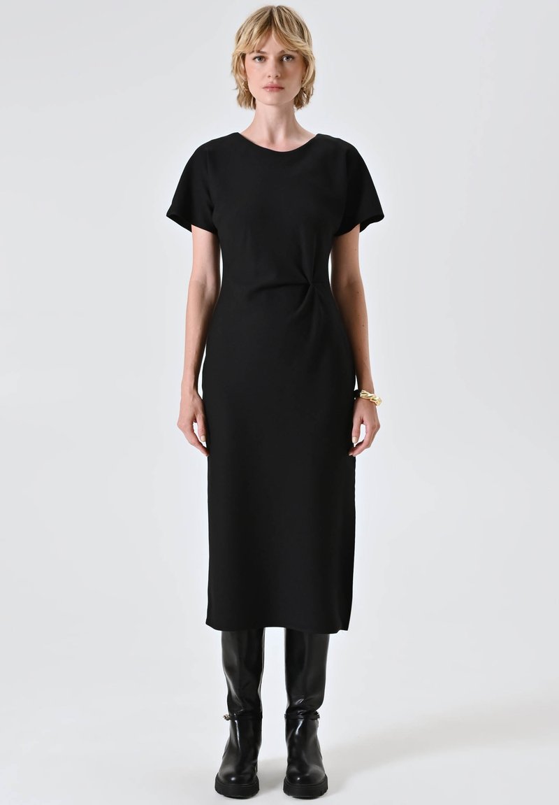 Abito nero midi con maniche corte, realizzato in un tessuto morbido. Presenta un sottile dettaglio a torsione in vita. Abbinato a stivali neri al ginocchio.