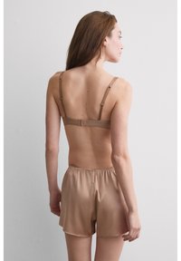 Kobieta o długich brązowych włosach, ubrana w beżowy biustonosz i pasujące satynowe szorty, stoi tyłem do prostej białej ściany.