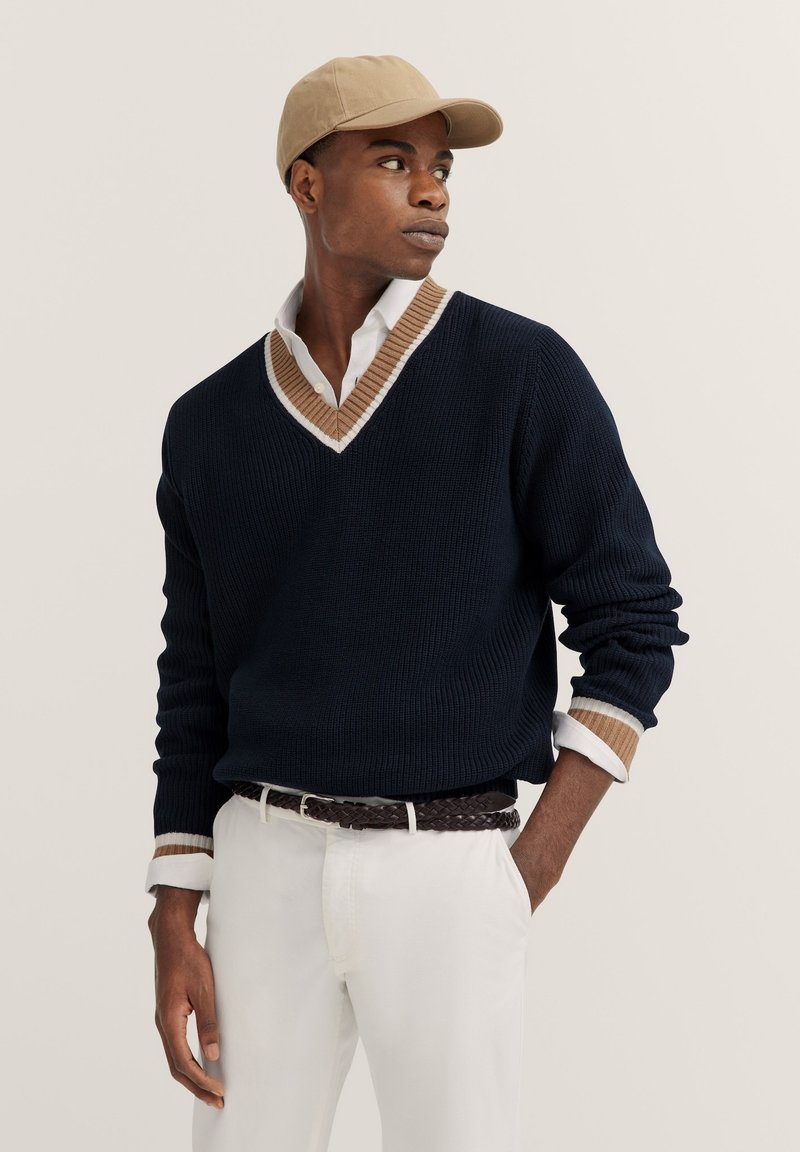 Dunkelblauer Strickpullover mit einem braunen und weißen gerippten V-Ausschnitt, darunter ein weißes Hemd, kombiniert mit weißen Hosen und einer beigen Mütze.