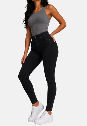 Femme portant un haut gris sans manches, un jean skinny noir taille haute et des baskets blanches, debout avec une main sur la hanche.