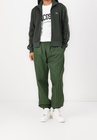 Chaqueta verde con cremallera y detalles en blanco, camiseta blanca con el logo de "Lacoste", pantalones verdes con estampado y zapatillas blancas. Conjunto deportivo casual.