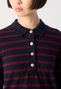 Young person wearing navy blue and burgundy striped polo shirt with four gold decorative buttons on placket.