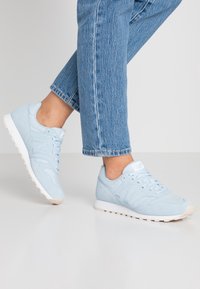 Des baskets en daim bleu clair présentent une semelle en caoutchouc blanc, des lacets et un logo sur le côté. Le design comprend un bout arrondi et un col rembourré.