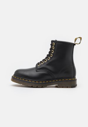 Schwarzer Leder-Schnürstiefel mit gelber Naht, dicker Gummisohle und einer gelb-schwarzen Zuglasche hinten auf schlichtem hellem Hintergrund.