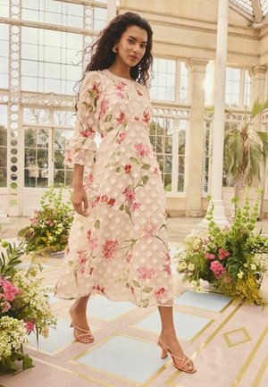 Frau geht drinnen und trägt ein weißes Midikleid mit pinken Blumenmustern und Rüschenärmeln, umgeben von bunten Blumenarrangements.