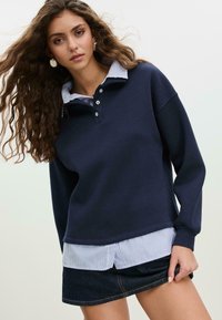 Marinblå huvtröja med ribbad krage över en blå och vit randig skjorta, tillsammans med en mörk denimkjol. Framträdande knappar på kragen.
