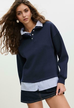 Felpa blu navy con colletto a costine sopra una camicia a righe blu e bianche, abbinata a una gonna di jeans scuri. Bottoni prominenti sul colletto.