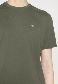 Olivengrøn bomuldst-shirt med rund hals og korte ærmer. Har et lille sølvbroderet logo på venstre bryst. Glat tekstur.