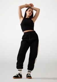 Crni rebrasti crop top, crne trenirke s bijelim bočnim prugama i crne tenisice s ružičastim detaljima. Model stoji s podignutim rukama.