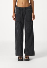 Bredde bukser i svart denim med elastisk midje, med to frontlommer og synlige sømmer. Brukt med svarte platåsandaler.
