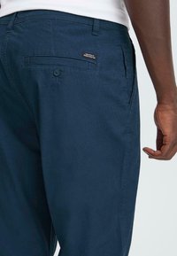 Moški oblečen v mornarsko modre hlače in belo srajco, ki prikazuje zadnji žep z gumbom in majhno črno nalepko blizu pasu.