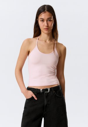 Jeune femme aux longs cheveux foncés portant un haut rose clair à col halter et un jean noir avec une ceinture, debout devant un fond blanc uni.