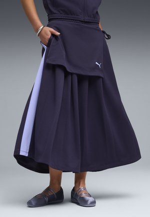 Personne portant une jupe évasée bleu marine avec une bande latérale violet clair, un haut assorti, un bracelet argenté et des chaussures à brides bleu marine.