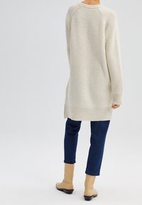 Maglione lavorato a maglia di un beige chiaro con una texture a costine, maniche lunghe e spacchi laterali, abbinato a jeans blu scuro e stivaletti beige.