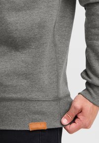 Pilkas flisinis džemperis su banguotomis rankogaliais ir apačia. Apačioje kairėje yra tekstūruotas ženklas su prekės pavadinimu.