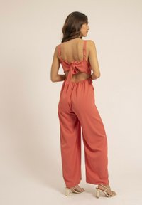 Jumpsuit corail à jambes larges, dos ouvert et bretelles ajustables. Présente un détail en nœud à l'arrière et une texture lisse et mate.