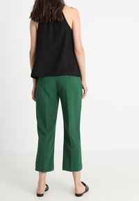 Black sleeveless top with a loose fit, paired with green cropped trousers. The outfit is completed with black flat sandals.