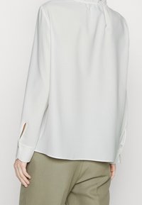 Blouse blanche à manches longues avec une coupe décontractée, dotée de poignets fendus et d'un col froncé. Tissu lisse, design minimaliste, sans motifs visibles.