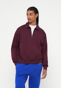 Bordeaux pullover med halv lynlås i kraven, lange ærmer og ribbede detaljer ved kanten. Bæret med blå svedbukser. Glat stoftekstur.