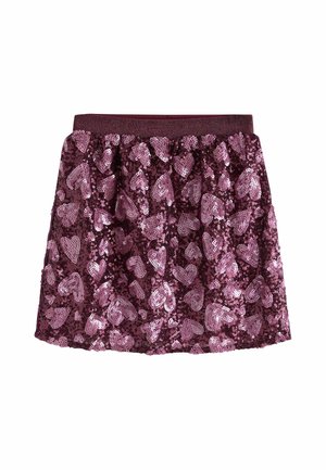 Jong meisje met gevlochten haar, een donker T-shirt, een roze rok met hartjesmotief en sneakers staat tussen rosegouden ballonnen op een effen achtergrond.