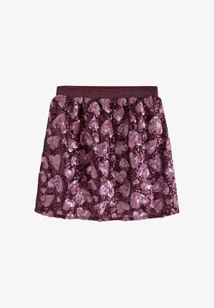 Junges Mädchen mit geflochtenem Haar, das ein dunkles T-Shirt, einen Rock mit pinkem Herzmuster und Turnschuhe trägt, steht zwischen rosegoldenen Luftballons vor einem schlichten Hintergrund.