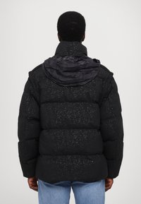 Black puffer jacket with a textured pattern, large collar, and a prominent hood. Features a boxy shape and thick insulation.