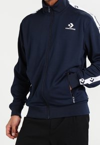 Chaqueta de chándal con cremallera de color marino, hecha de un tejido suave; cuenta con rayas blancas y un logo de estrella a lo largo de las mangas, además de bolsillos con cremallera y un cuello alto.