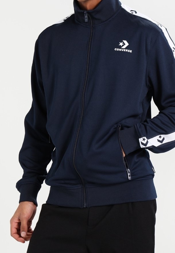 Chaqueta de chándal con cremallera de color marino, hecha de un tejido suave; cuenta con rayas blancas y un logo de estrella a lo largo de las mangas, además de bolsillos con cremallera y un cuello alto.