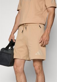 Beige athletic shorts with drawstring waist, side pockets, and a subtle three-stripe accent on the leg. Material appears soft and flexible.