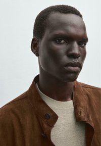 Veste en daim marron avec des accents de boutons sur un pull en tricot beige clair. Gros plan sur les détails sur un fond neutre.