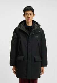 Giacca parka imbottita nera con cappuccio, dotata di zip frontale e bottoni a pressione, due tasche frontali e un logo discreto sul petto.