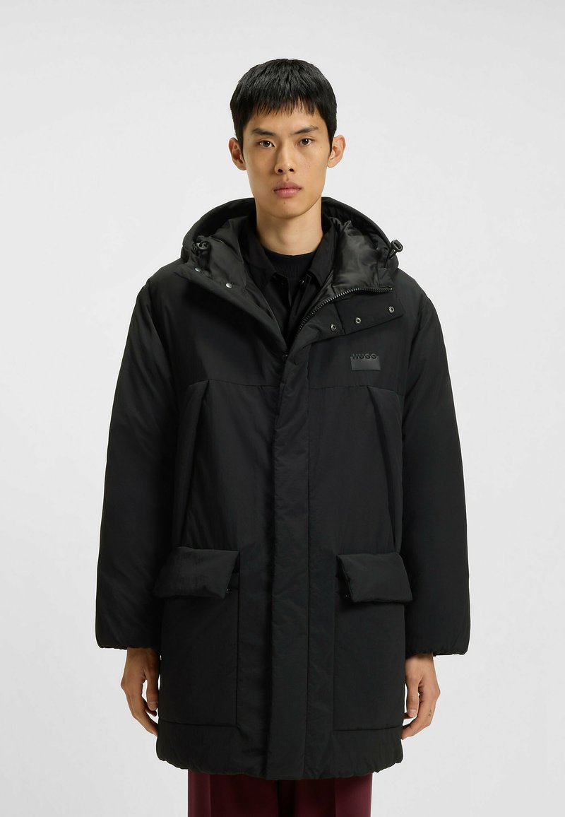 Zwarte gewatteerde parka met capuchon, voorzien van een rits aan de voorkant en drukknoopjes, twee voorzakken en een subtiele logo op de borst.
