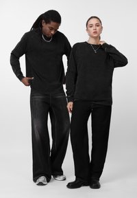 Dos personas de pie una al lado de la otra, con camisetas negras de manga larga y pantalones negros de pierna ancha, una mirando hacia abajo y la otra hacia adelante.