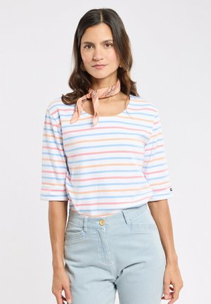 Femme portant une chemise à rayures pastel avec des manches jusqu'au coude, un pantalon à fines rayures bleu clair et un foulard pêche autour du cou, sur fond blanc.