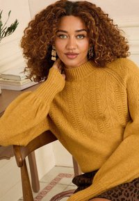 Mustár sárga kötött pulóver magas gallérral, texturált mintával és bordás mandzsettával. Lazább fazon. Sötét mintás szoknyával viselve.