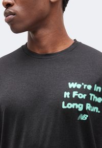 Gros plan d'une personne portant un t-shirt gris foncé avec un texte bleu clair disant "We're In It For The Long Run" et un logo New Balance.