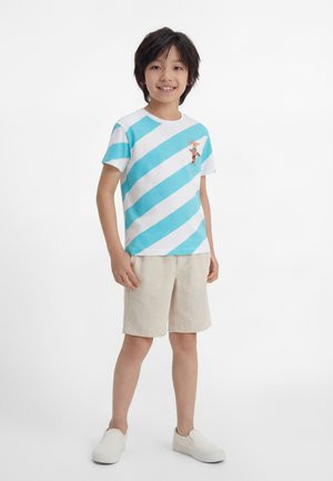 Băiat stând zâmbind, purtând un tricou cu dungi diagonale albastre și albe cu un desen de desene animate, pantaloni scurți bej și pantofi slip-on albi.