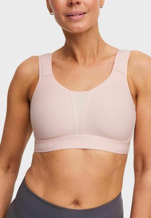 Vrouw draagt een lichtroze sportbeha met brede bandjes en een detail van mesh panelen, gecombineerd met donkergrijze sportlegging.