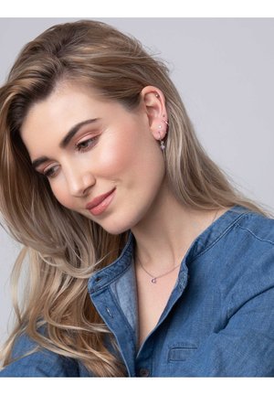 Zilveren sieraden met een hanger met een paarse edelsteen. Model draagt een denim shirt. Zachte golven in het haar, met meerdere oorpiercingen zichtbaar.