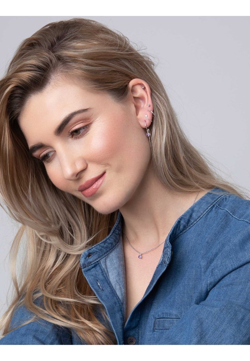 Zilveren sieraden met een hanger met een paarse edelsteen. Model draagt een denim shirt. Zachte golven in het haar, met meerdere oorpiercingen zichtbaar.