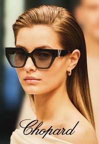 Femme aux cheveux tirés en arrière, portant de grandes lunettes de soleil noires de créateur et de petites boucles d'oreilles en or en forme d'anneau, avec le texte "Chopard" en bas.