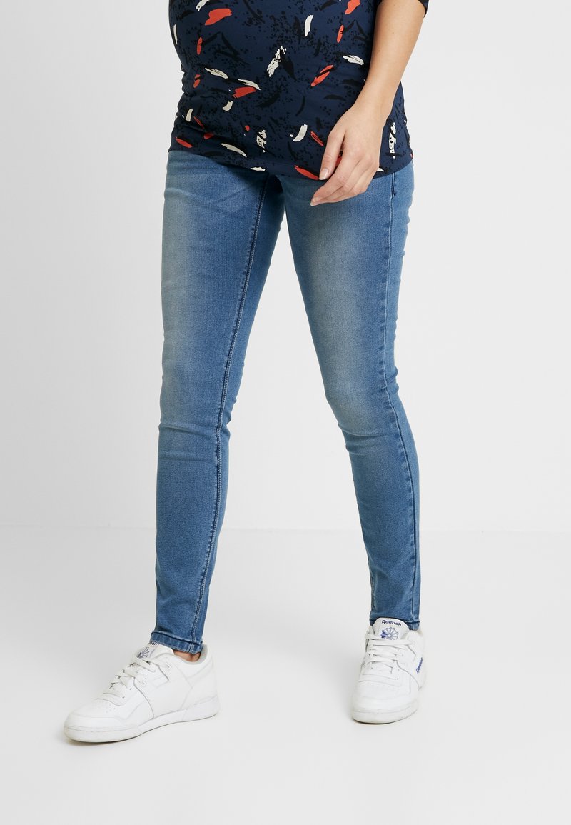 Jeans skinny azul com uma textura ligeiramente desbotada e costuras em contraste, combinados com ténis brancos. Sem padrões visíveis ou desgastes.