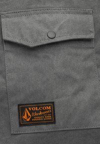 Grahamare Stofftasche mit einem schwarzen Druckknopf und einem orangefarbenen Volcom Workwear Etikett, das "Qualität und haltbare Waren" sagt.