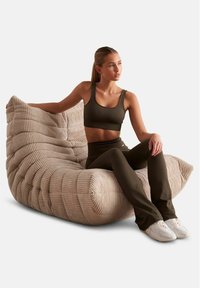 Chaise longue beige à rayures avec un design sculpté et courbé. Une personne en tenue de sport olive est assise de manière décontractée, mettant en valeur la texture luxueuse de la chaise.