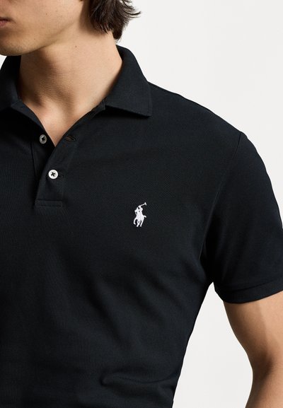 Sort polo-shirt lavet af struktureret stof, med en tre-knaps placket og et hvidt broderet logo på venstre bryst.