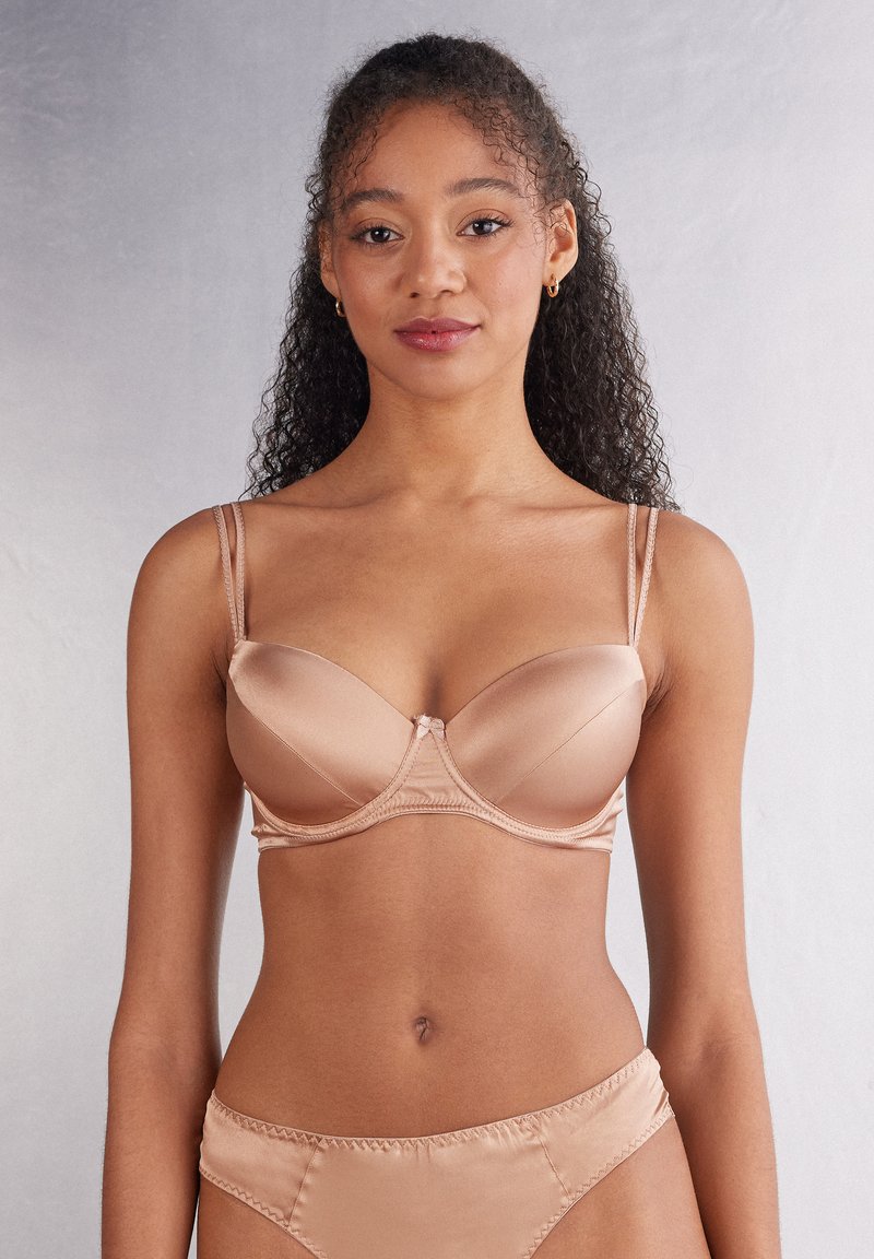 Nahá saténová podprsenka s drátem, nastavitelnými ramínky a centrální mašlí. Slaďena s odpovídajícími saténovými kalhotkami.