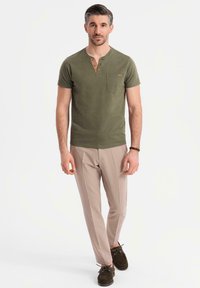 Olivově zelená krátká rukávová košile Henley s knoflíkovou légou a kapsou, v kombinaci se světle béžovými kalhotami a tmavě hnědými botami.