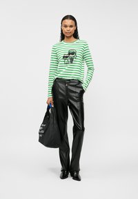 Zöld és fehér csíkos, hosszú ujjú ing grafikai nyomattal, fekete bőrnadrággal párosítva. A modellen fekete vászontáska van.