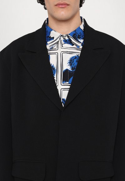 Black tailored blazer with wide lapels, paired with a colorful, patterned blue and white shirt featuring graphic designs.